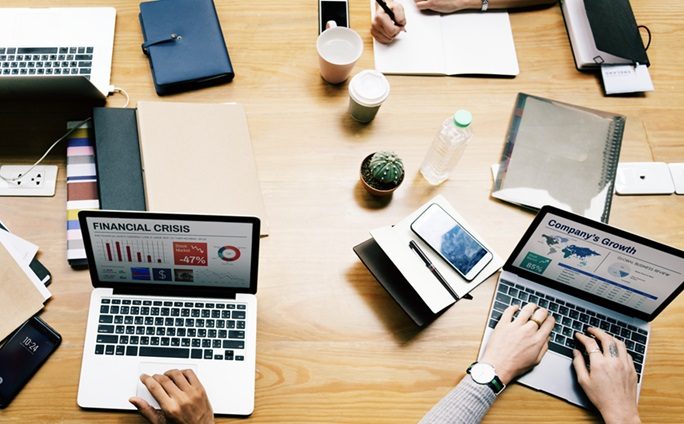 Colleagues working on laptops around a shared desk with business dashboards on screen